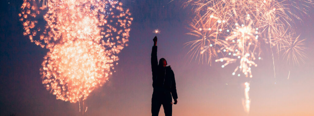 Eine Person steht in der Abenddämmerung im Freien und hält eine brennende Wunderkerze in die Höhe, während hinter ihr große bunte Feuerwerkskörper am Himmel explodieren.